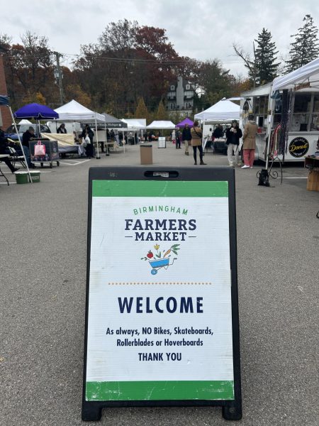 This welcome sign can be seen at the entrance of the Birmingham Farmers Market, hosted in parking lot number 6 in the heart of downtown Birmingham. In the background, vendors are standing at their stalls, and a group of people can be seen congregated near one of the several food trucks parked around the market.