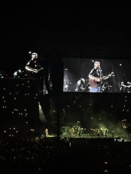 Zach Bryan projected onto one of the numerous televisions surrounding the center of University of Michigan's Big House stadium. In the dark stadium, the onlookers flashlights shine and the stage illuminates by a dim yellow.
