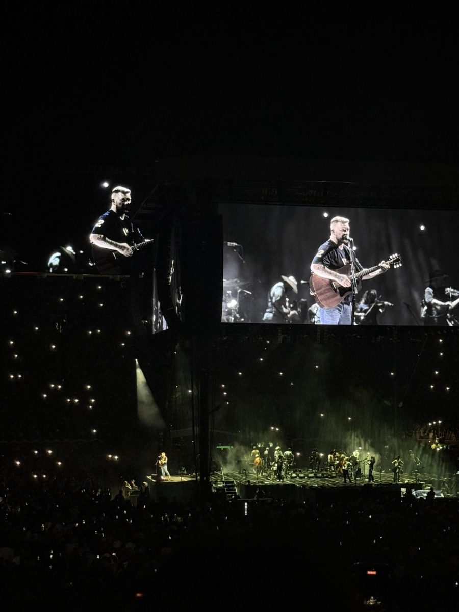 Zach Bryan projected onto one of the numerous televisions surrounding the center of University of Michigan's Big House stadium. In the dark stadium, the onlookers flashlights shine and the stage illuminates by a dim yellow.