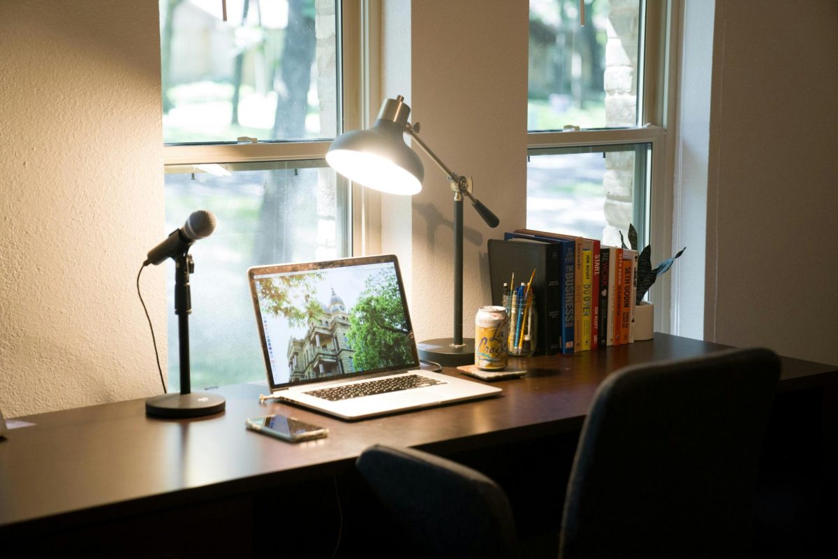 Photo with a desk that has a laptop on top of it, with books, and lamp, and more. This demonstrates a typical space for a student to be studying at. (Source via Unsplash.)
